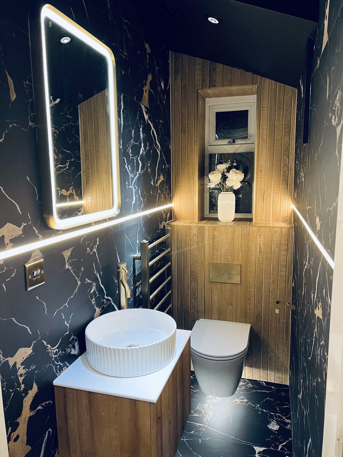 Cloakroom with black and gold marble walls, a fluted vessel basin and a backlit mirror