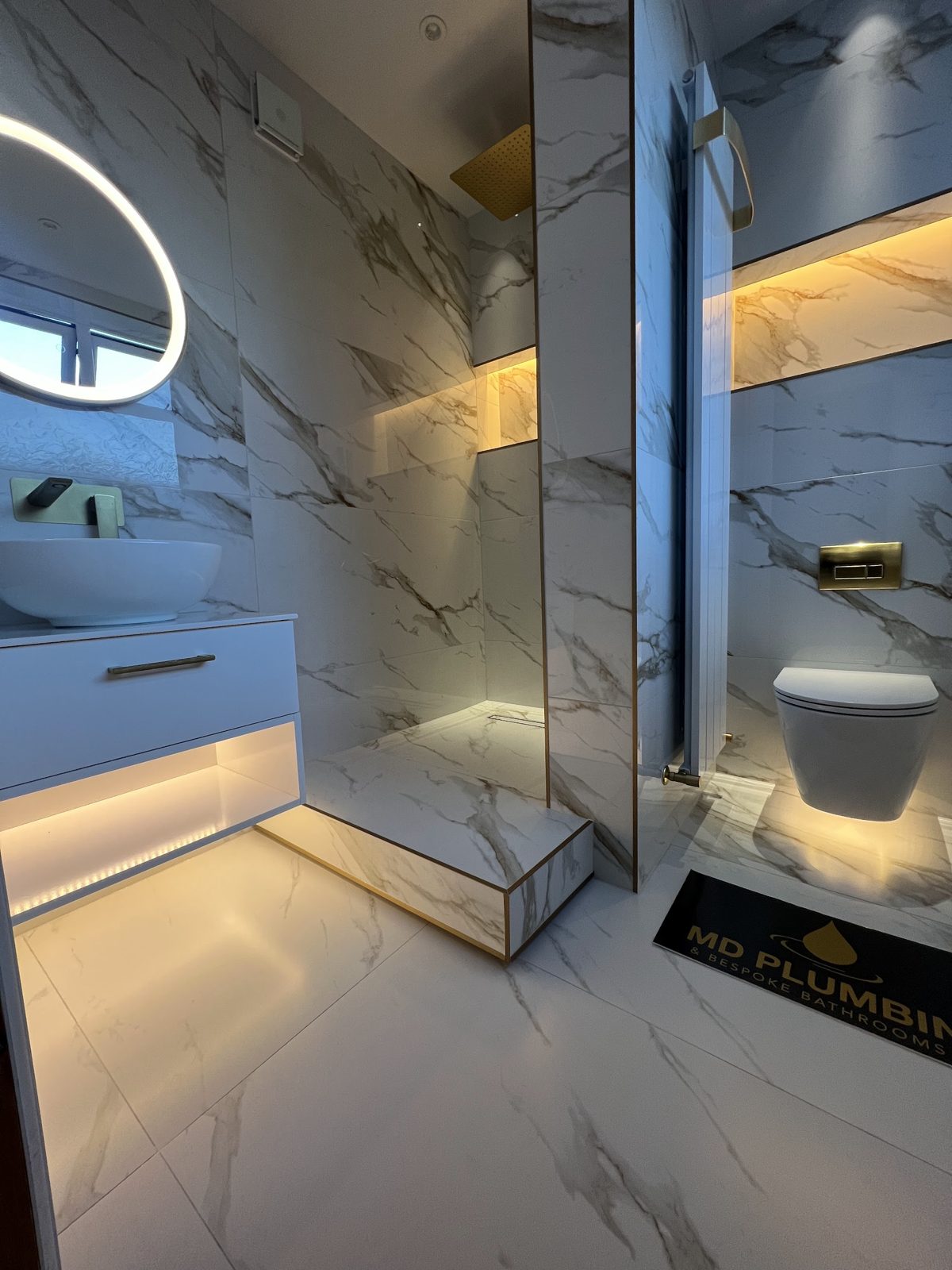 White marble bathroom with a lit plinth and brushed brass flush plate