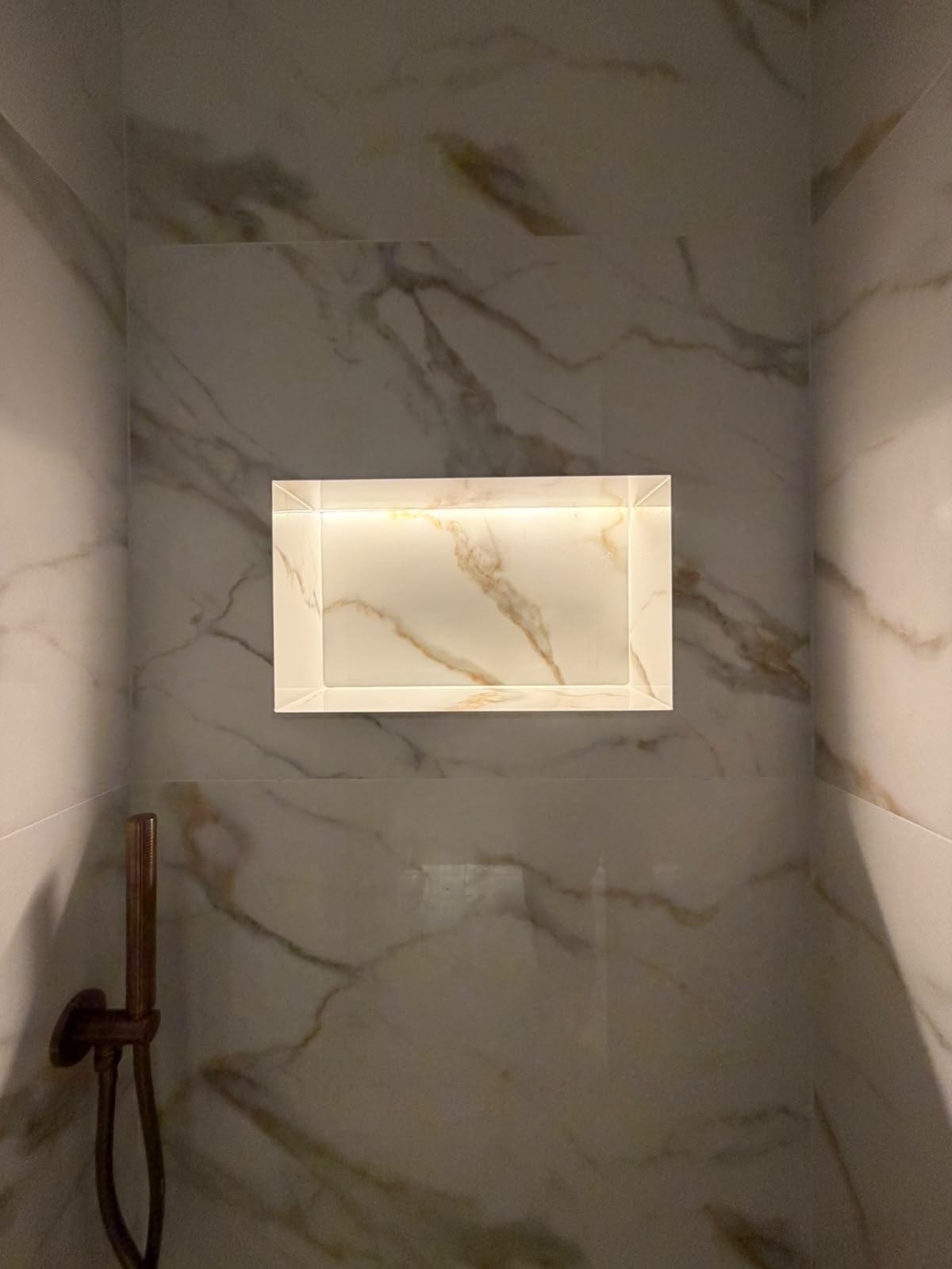 Gold-veined marble niche lit from within beside a brushed bronze shower